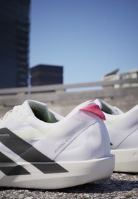 Paar weiße Adidas Adizero Adios 9 Laufschuhe mit schwarzen Streifen und pinkfarbenen Fersenlaschen auf einem Außenpflaster unter klarem Himmel.