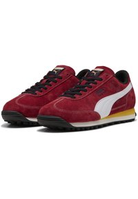 Adidași atletici din suede roșu cu accente albe, șireturi negre și talpă din cauciuc, având un toc galben și detalii cu logo pe limbă.