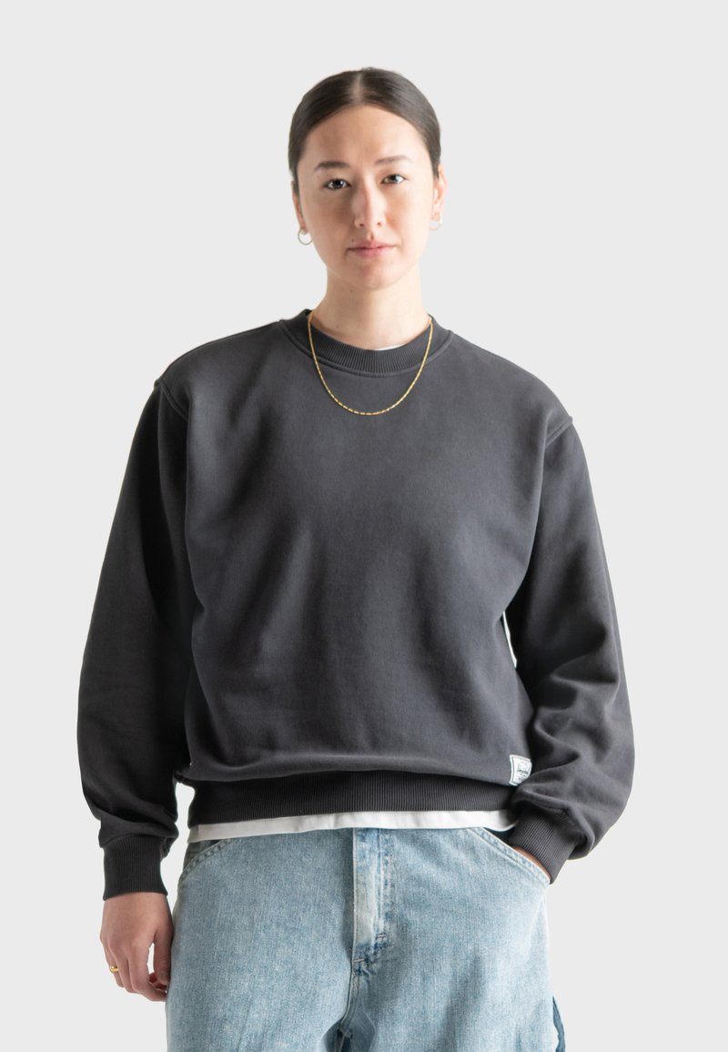 Sudadera de color gris oscuro con cuello redondo y puños acanalados, combinada con jeans de color azul claro. Presenta una etiqueta de logo sutil en el dobladillo.