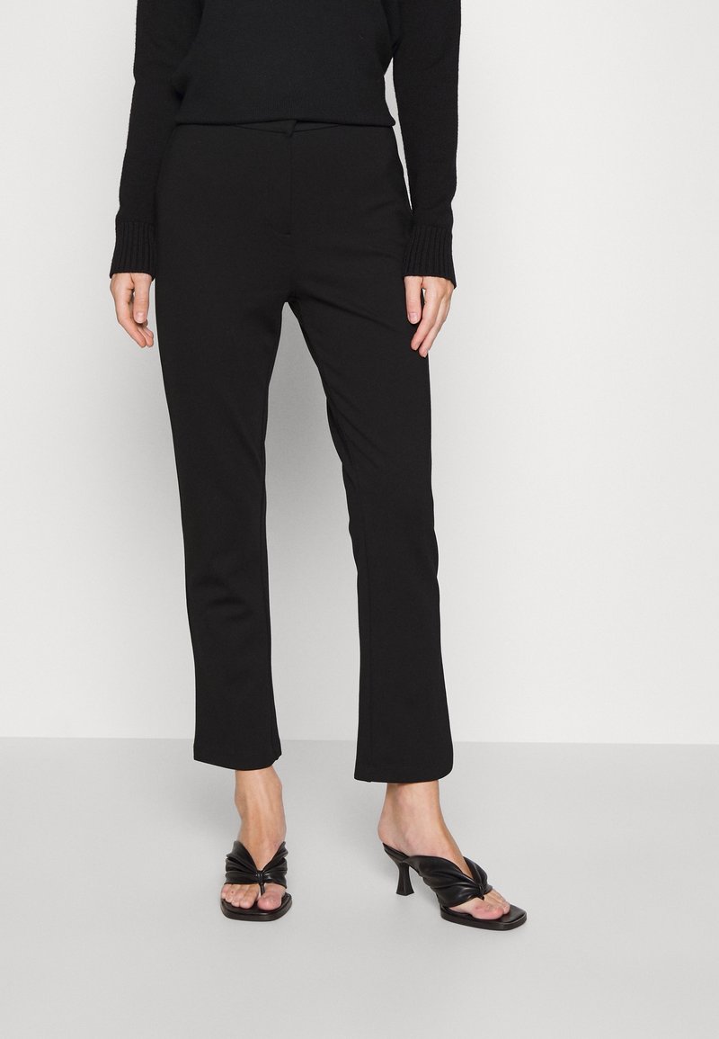 Femme portant un pantalon droit noir, un pull à manches longues noir et des sandales à talons noires, debout devant un fond blanc uni.