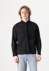 Black textured long-sleeve shirt with raised dot pattern, band collar, and front pocket, paired with light blue jeans.