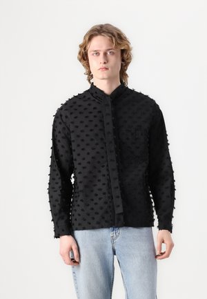 Maglietta nera a maniche lunghe con texture e motivo a puntini in rilievo, colletto a band, e taschino frontale, abbinata a jeans azzurri chiari.