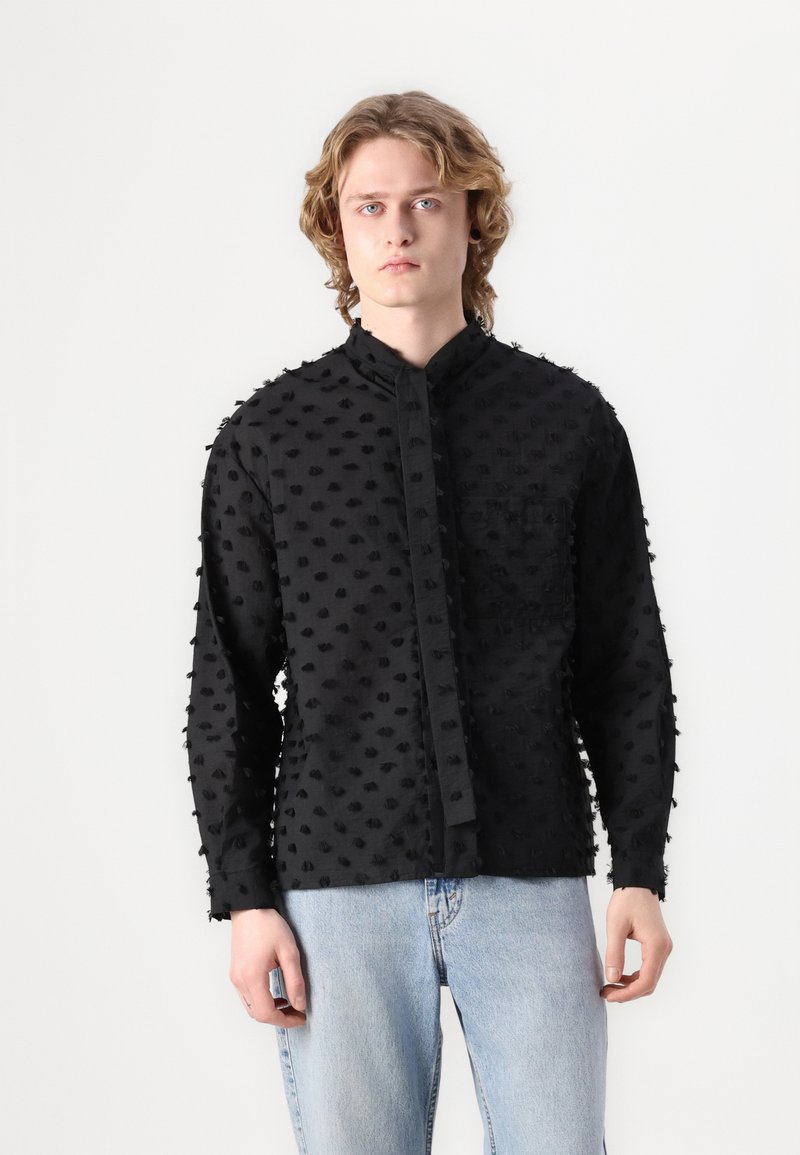 Black textured long-sleeve shirt with raised dot pattern, band collar, and front pocket, paired with light blue jeans.