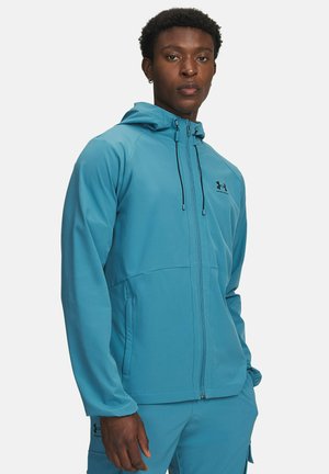 Lichte jas met capuchon in levendig teal, voorzien van een rits aan de voorkant, verstelbare trekkoorden en zijzakken. Zwart logo op de borst.