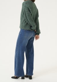 Grüner gestrickter Pullover mit strukturierter Oberfläche, kombiniert mit hoch taillierter blauer Jeans im Wide-Leg-Stil und schwarzen Lackleder-Pumps mit Blockabsatz.