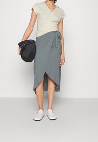 Light yellow top with a v-neck and gathered sides, paired with a gray wrap skirt featuring a tie and uneven hem. Wearing pale shoes and holding a black bag.