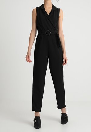 Frau trägt ärmellosen schwarzen Overall mit Gürtel und weitem Kragen sowie schwarze Schuhe mit runden Schnallendetails.