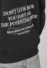 Sweatshirt noir avec texte blanc : « NE REGARDE PAS EN ARRIÈRE, TU AS TOUT LE POTENTIEL À L'INTÉRIEUR » avec des accents graphiques. Texture douce, poignets et ourlet côtelés.