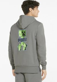 Grå hættetrøje med et stort grønt Minecraft Creeper-tryk på ryggen, samt "PUMA" tekst nedenfor. Glat stof, standard pasform, snørret hætte.