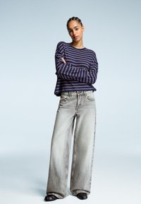 Navy striped long-sleeve top with red lines; loose-fitting gray wide-leg jeans; black flat shoes; minimalistic background.