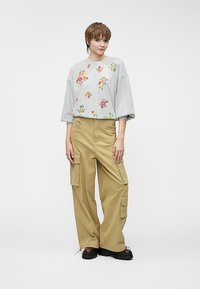 Šedá mikina s kvetinovou výšivkou; voľný strih, okrúhly výstrih. Voľné béžové cargo nohavice s bočnými vreckami, nosené s robustnými čiernymi topánkami.