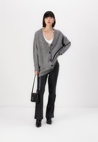 Cárdigan gris de gran tamaño con botones, llevado sobre una blusa blanca. El modelo lo combina con pantalones de cuero negros y sostiene un pequeño bolso negro.