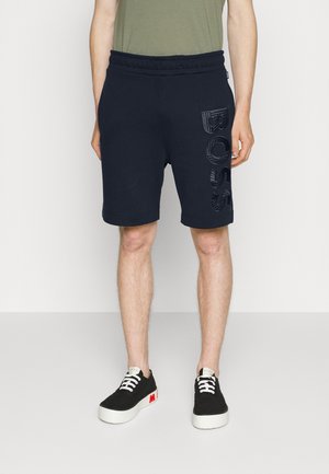 Homme portant un short noir avec un grand lettrage en relief "BOSS" sur la cuisse gauche, assorti à des baskets noires et blanches.