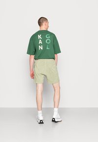 Groen oversized t-shirt met grote, meerkleurige letters "KAN GOL" op de achterkant. Lichtgroene badstof shorts. Witte hoge schoenen.