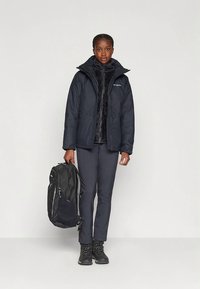 Casaco acolchoado preto com gola alta, usado sobre um top de fleece preto. Calças escuras e botas. Segurando uma mochila preta com detalhes em cinza.