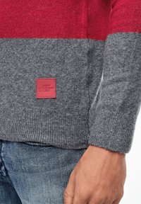 Homme portant un pull en maille rouge et gris avec une pièce rouge, associé à un jean en denim bleu, la main détendue sur le côté.