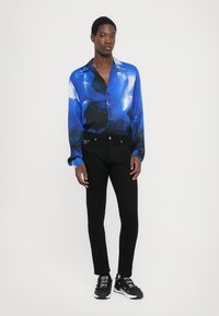 Camicia button-up blu e nera con motivo, texture setosa, maniche lunghe, abbinata a jeans skinny neri e sneakers nere.