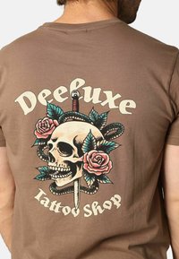 Bruine katoenen t-shirt met een grafisch ontwerp van een schedel met een dolk, omringd door rozen en bladeren. Tekst: "Deeluxe Tattoo Shop."