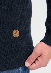 Dunkelblauer, texturierter Strickpullover mit Rippenmuster, versehen mit einem runden braunen Lederetikett an der Seite. Eine Hand greift den Saum.