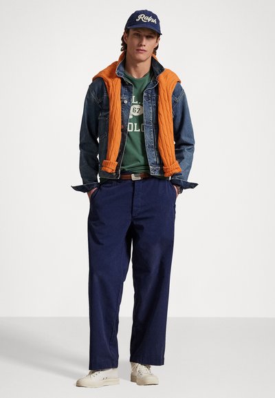 Veste en denim, t-shirt vert, pull orange drapé sur les épaules, pantalon bleu marine et baskets crème. Casquette avec logo blanc sur le devant.
