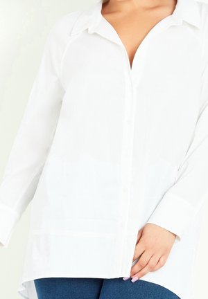 Biała koszula na długi rękaw z kołnierzykiem, noszona na niebieskich dżinsach, dłoń spoczywająca blisko biodra na krawędzi koszuli.