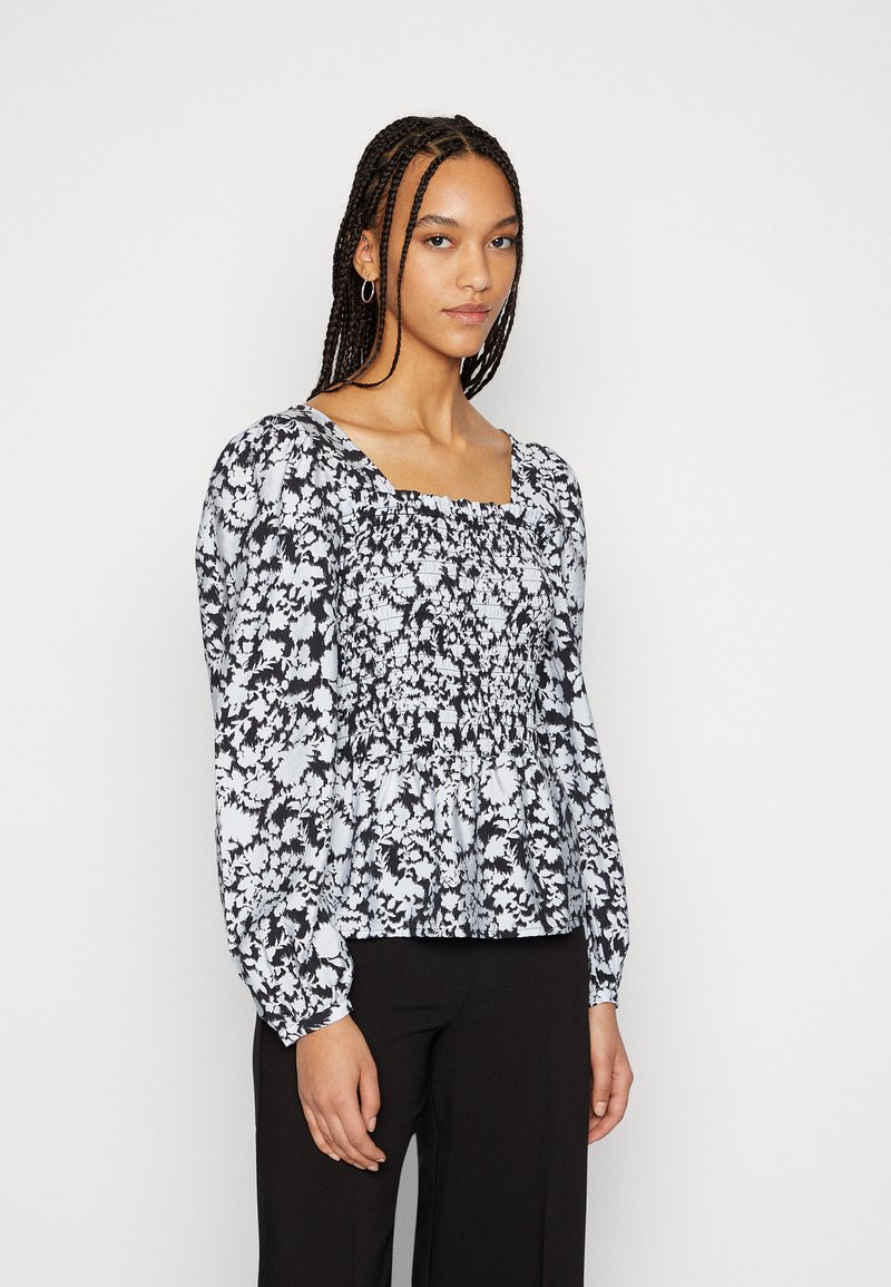 Blusa floral em preto e branco com mangas longas, decote quadrado e detalhes franzidos na cintura, combinada com calças pretas.