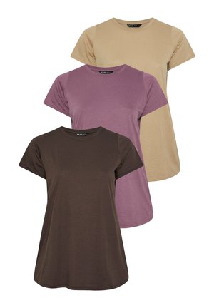 Donna bionda in piedi contro un muro, indossa una maglietta viola ampia, jeans larghi celeste chiaro e sneakers bianche.