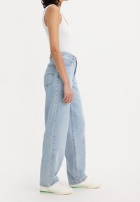 Femme portant un jean large bleu clair, un débardeur blanc et des baskets blanches, debout de profil sur un fond blanc.