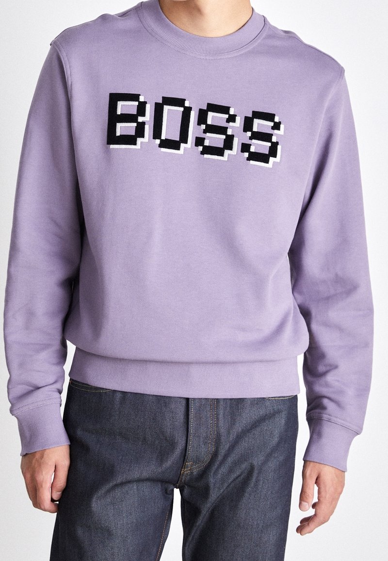 Personne portant un sweat-shirt violet clair avec un texte pixellisé en noir et blanc "BOSS" sur la poitrine et un jean bleu foncé.