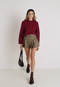 Bordový pletený sveter s golierom, v kombinácii s kockovanou plisovanou sukňou. Modelka má na sebe hnedé ponožky a čierne platformové topánky, v ruke drží malú tašku.