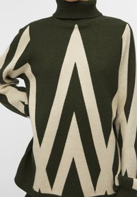 Groene en beige gebreide trui met een hoge nek, met een zigzagpatroon. Het ontwerp heeft een losvallende pasvorm en ribgebreide manchetten.