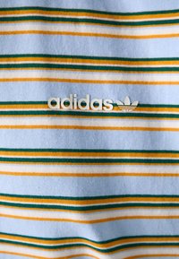Tissu bleu clair avec des rayures horizontales vertes, orange et blanches, avec un logo adidas blanc et un symbole trefoil au centre.