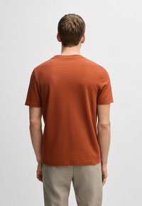T-shirt arancione ruggine a maniche corte realizzato in tessuto testurizzato con scollatura rotonda e vestibilità rilassata, abbinato a pantaloni chiari.