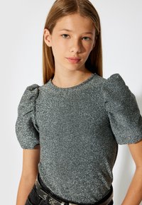 Glitzerndes graues Oberteil mit kurzen Puffärmeln. Glatte Textur und taillierte Passform. Kombiniert mit einem schwarzen Gürtel mit silbernen Akzenten.