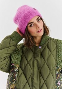 Růžová pletená čepice s texturovaným žebrovaným vzorem, kombinovaná se zelenou prošívanou bomber bundou s květinovým potiskem a akcenty z copánkového pletení.