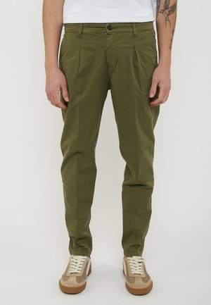 Uomo che indossa pantaloni chino verde oliva e sneakers beige, in piedi davanti a uno sfondo bianco semplice.