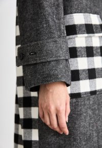 Sivý a čierny károvaný kabát s houndstooth vzorom. Textúrovaná vlnená látka, kontrastný detail na vrecku a gombíkový náplet.