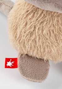 Plush toy with a beige fluffy body, gray limbs, and a red tag featuring a white paint splatter. Soft texture and rounded shapes.