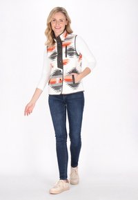 Gilet a fantasia in crema, arancio e nero con zip frontale, tasche e colletto alto, indossato sopra una maglietta bianca a maniche lunghe.