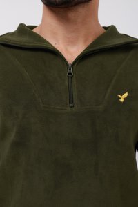 Olivengrøn fleece-pullover med kvart lynlås og blød tekstur. Har et lille gult fugle-emblem på brystet.