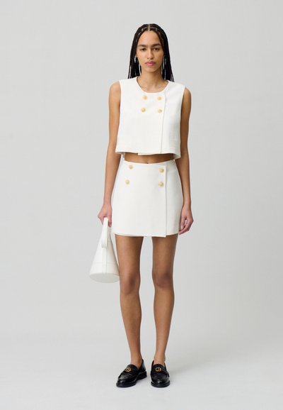 Haut court blanc avec des boutons dorés décoratifs et une jupe courte, tous deux en tissu lisse. Modèle tenant un sac à main blanc et portant des mocassins noirs.