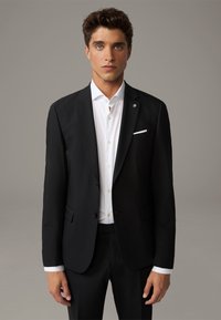 Schwarze Anzugjacke mit schlanker Passform, zwei Knöpfen und Revers. Sie verfügt über ein weißes Taschentuch und ein strukturiertes Gewebe. Kombiniert mit einem weißen Hemd mit Knopfleiste.