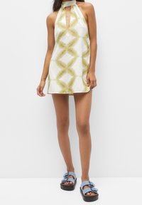 Donna che indossa un miniabito senza maniche con scollo a halter, caratterizzato da un motivo geometrico giallo e bianco, e sandali con piattaforma blu chiaro.