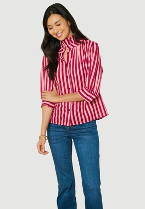 Femme aux longs cheveux foncés souriante, portant une blouse à rayures verticales rouges et roses et un jean bleu, debout devant un fond clair uni.