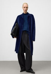 Frau in einem dunkelblauen, flauschigen Mantel über einem blauen Pullover, schwarzen, weitgeschnittenen Hosen und spitzen schwarzen Schuhen, die eine große schwarze Ledertasche hält.