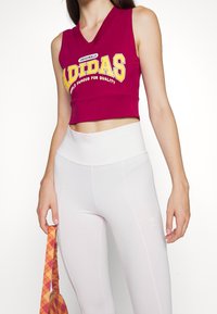 Burgundská bezramínková crop top s žlutým logem "Adidas", spárovaná s bílými legínami s vysokým pasem a diskrétním detailem loga.