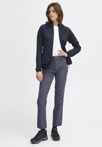 Giacca nera con zip e cappuccio, t-shirt bianca, pantaloni grigi e scarpe da ginnastica nere. Tessuto liscio, vestibilità su misura e chiusura con due bottoni sui pantaloni.