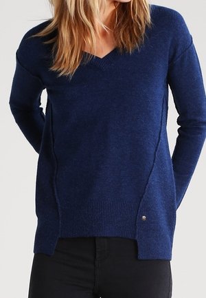 Femme portant un pull à manches longues bleu marine avec un col en V, associé à un pantalon noir, debout les mains derrière le dos.