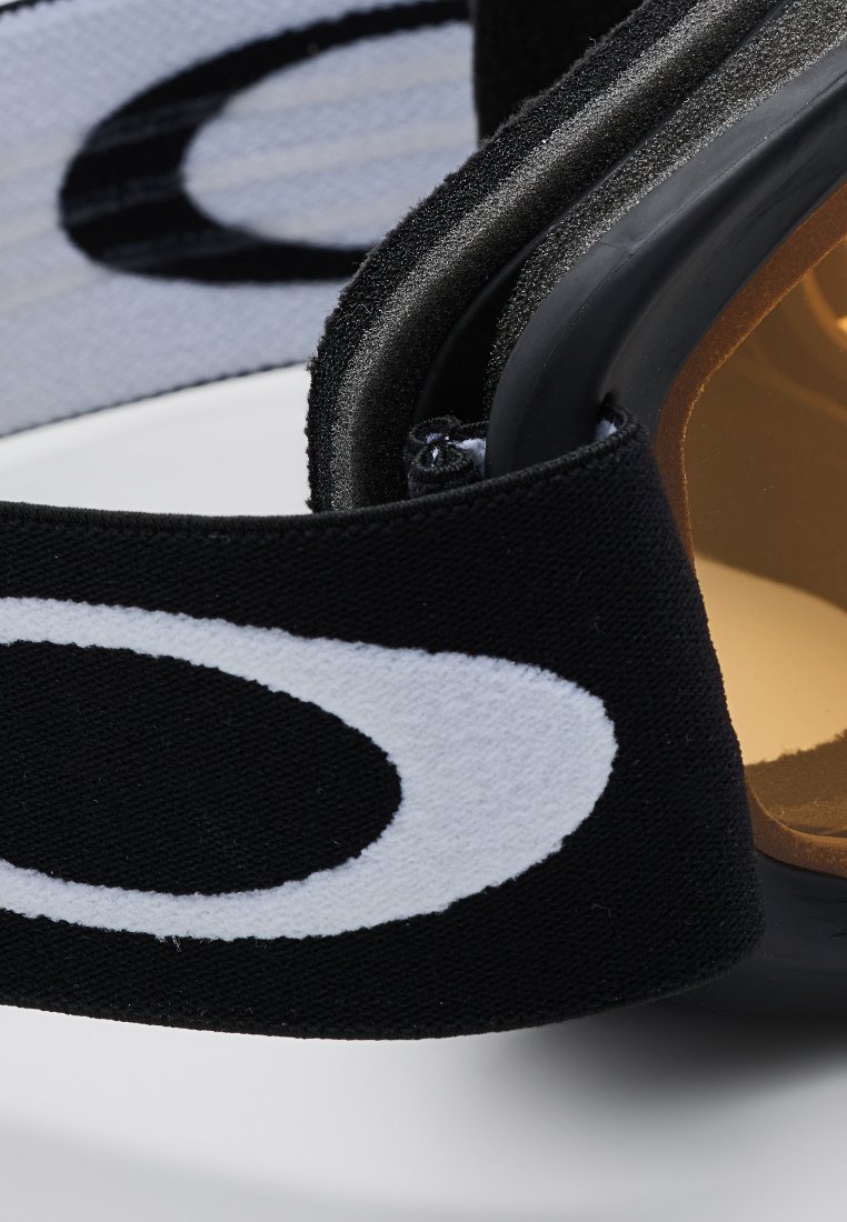 Zbliżenie czarnego pasku tkaninowego z białym owalnym logo przymocowanego do sportowych lub ochronnych okularów w odcieniu brązowym, z piankowym wykończeniem.