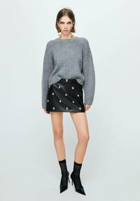 Jersey gris de punto, falda mini de piel sintética negra con tachuelas metálicas, medias negras transparentes y tacones altos de punta. Diseño simple y moderno.